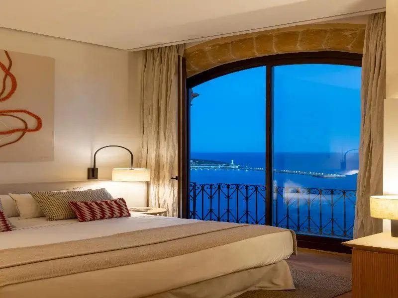 Schlafzimmer mit Doppelbett und Blick durch ein rundbogiges Fenster auf das Meer bei Dämmerung.
