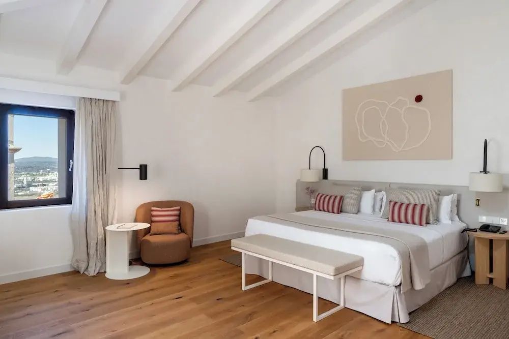 Helles Schlafzimmer mit weißer Holzbalkendecke, großem Doppelbett und einem Sessel in der Ecke.