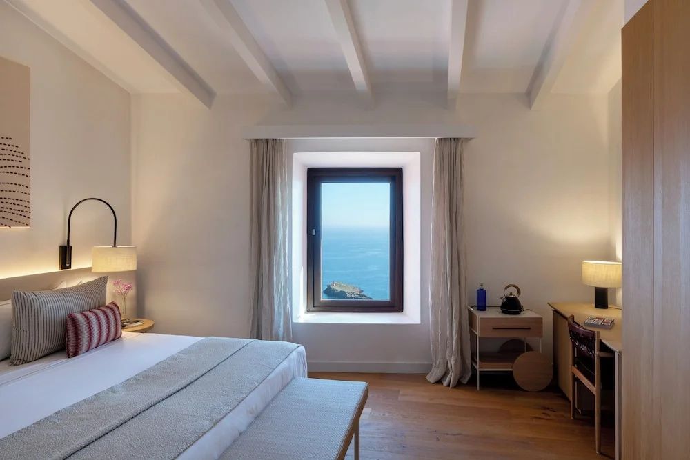 Helles Hotelzimmer mit Holzboden, weißer Holzbalkendecke und Fenster mit Blick auf das Meer.