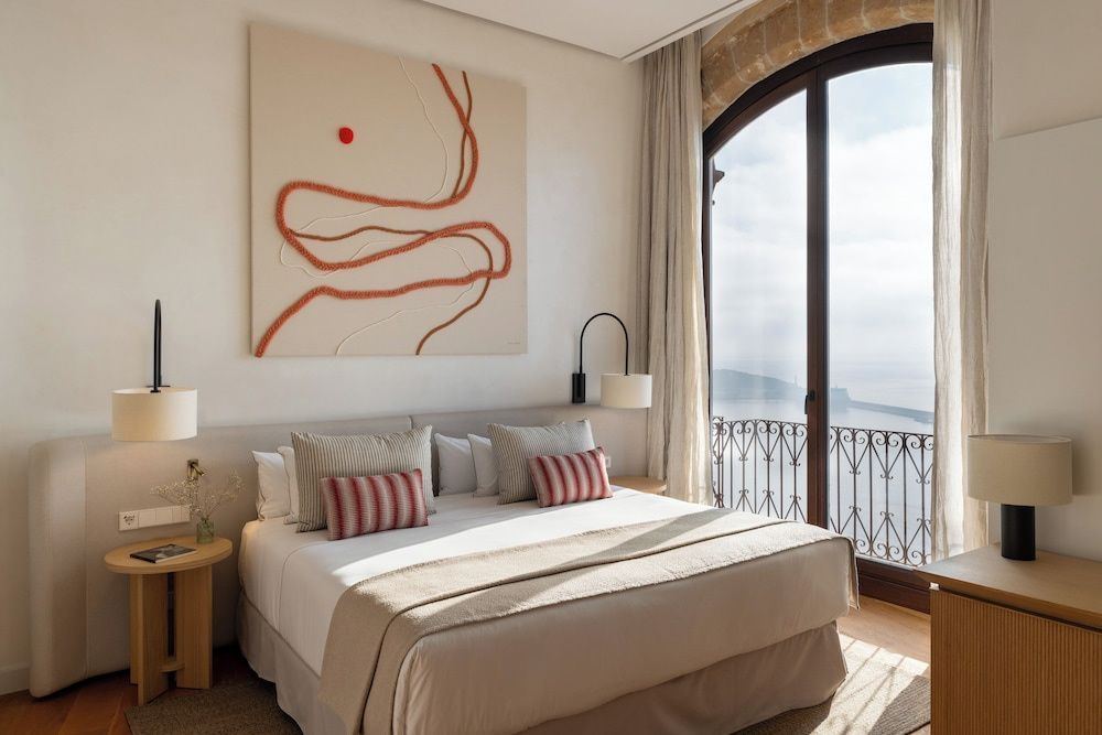 Modernes Schlafzimmer mit Doppelbett, Wandbild und hohem Bogenfenster zum Meer.
