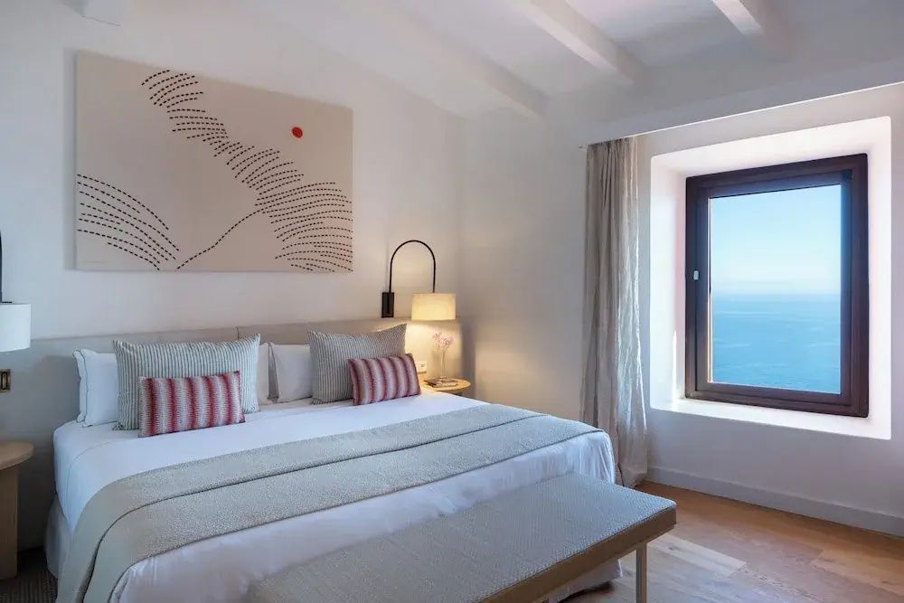 Helles Hotelzimmer mit Doppelbett, Wandbild und Fenster mit Ausblick auf das Meer.