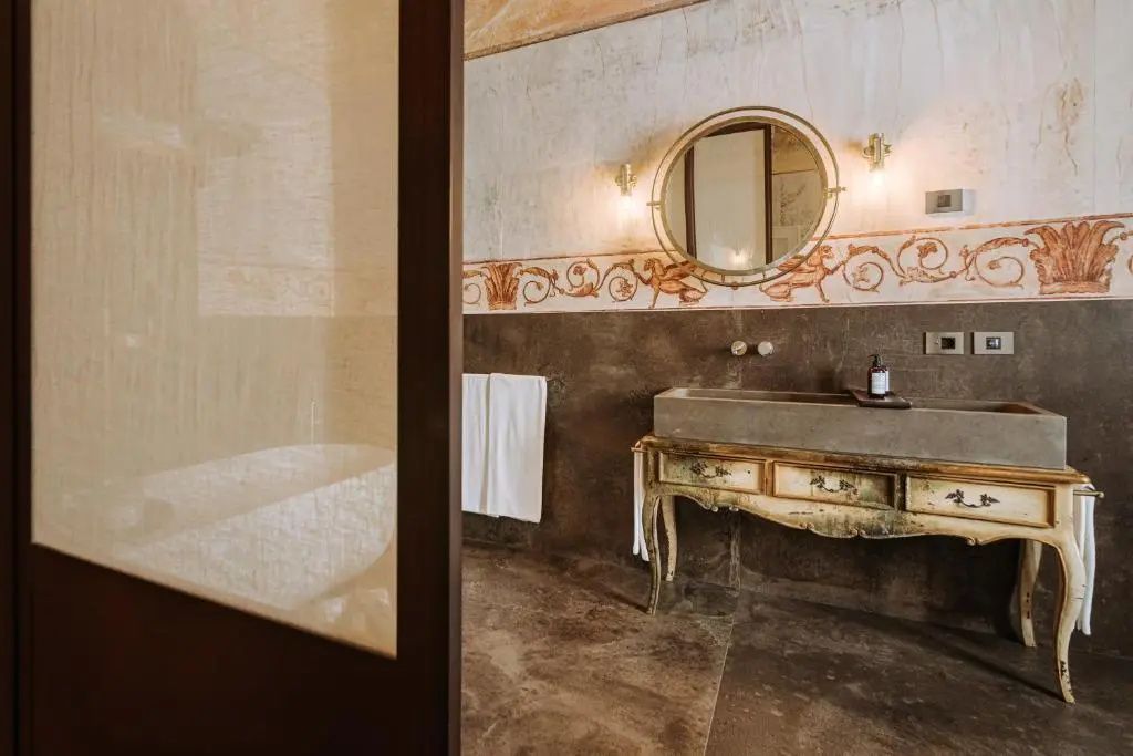Badezimmer mit Steinwaschbecken auf antikem Holztisch, rundem Spiegel und Blick zur Wanne.