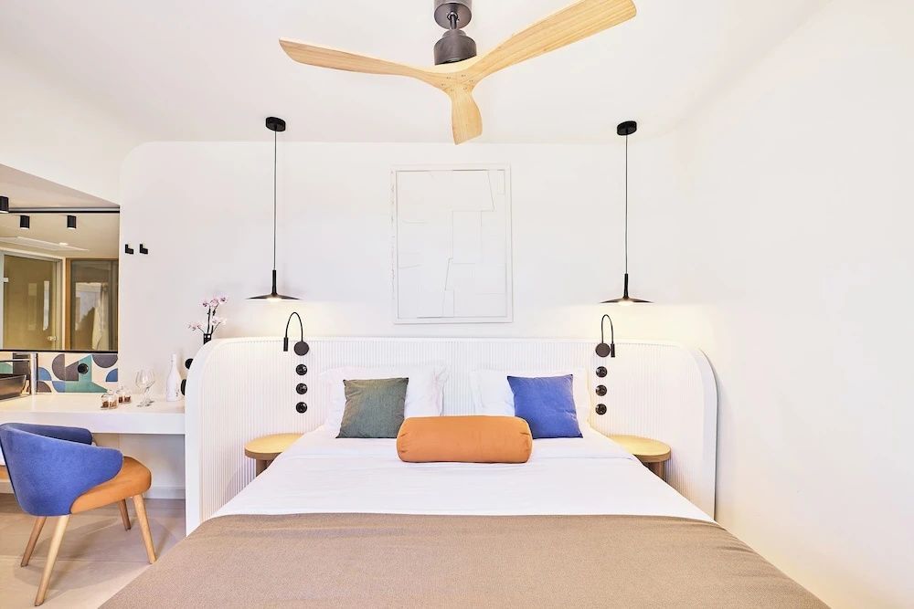 Helles Doppelbett mit bunten Kissen und einem Deckenventilator aus Holz.