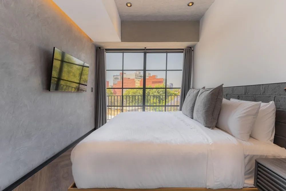 Doppelbett in modernem Hotelzimmer mit bodentiefem Fenster und Fernseher an der Wand.