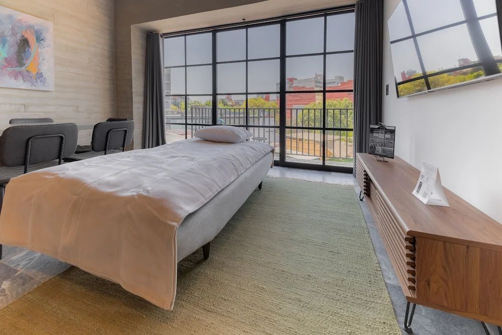 Einzelbett auf hellem Teppich in einem Hotelzimmer vor einer großen Fensterfront.