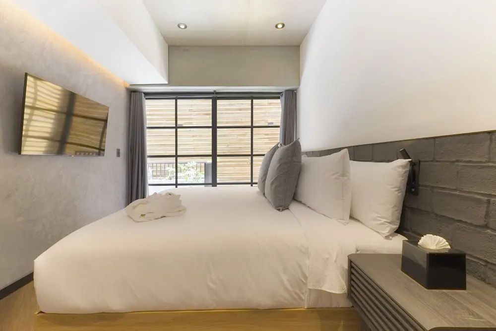Doppelbett mit weißer Bettwäsche, grauen Kissen und einem großen Fenster im Hintergrund.