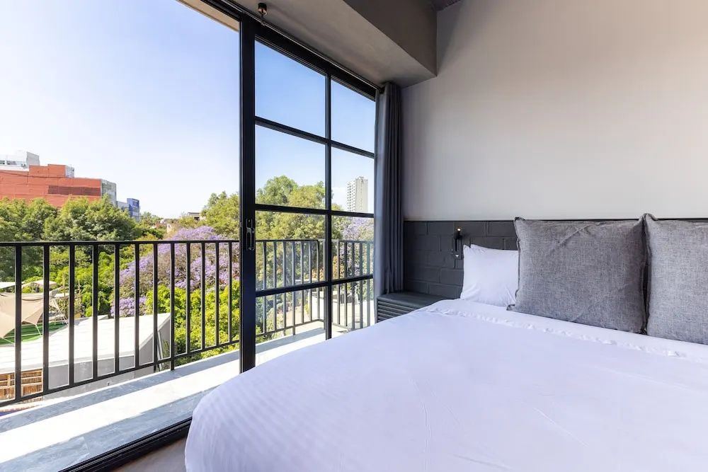 Bett im Schlafbereich mit großem Fenster und Ausblick auf grün bewachsene Bäume.