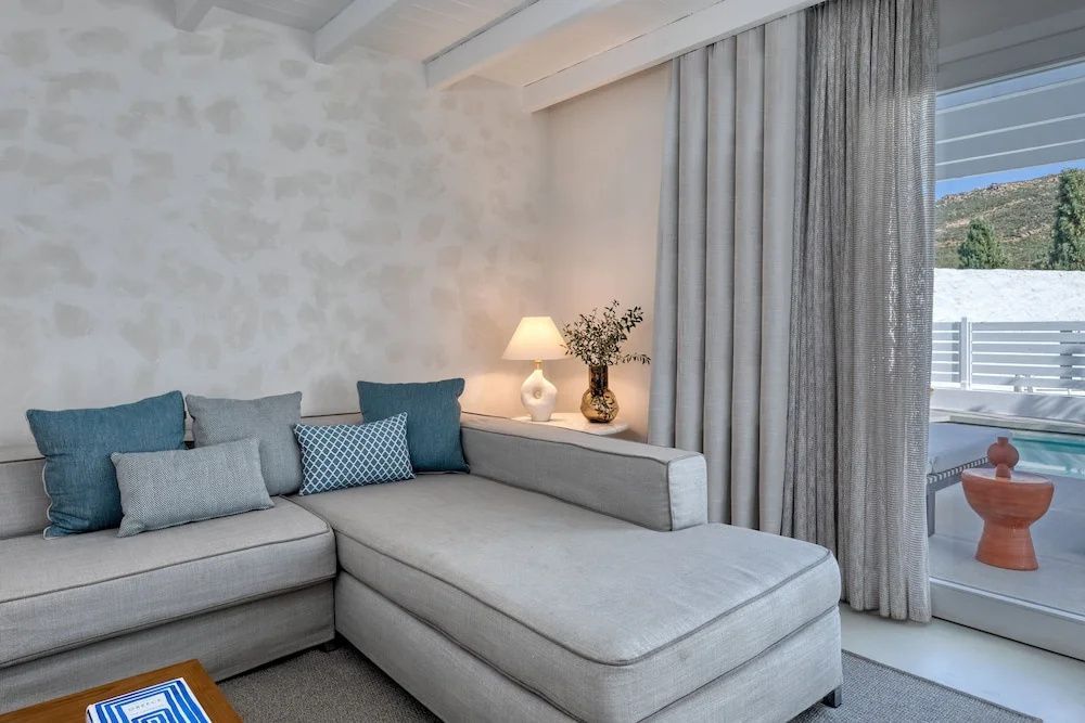 Sitzecke im Hotelzimmer mit hellgrauem Ecksofa, blauen Kissen und einer Tischlampe.