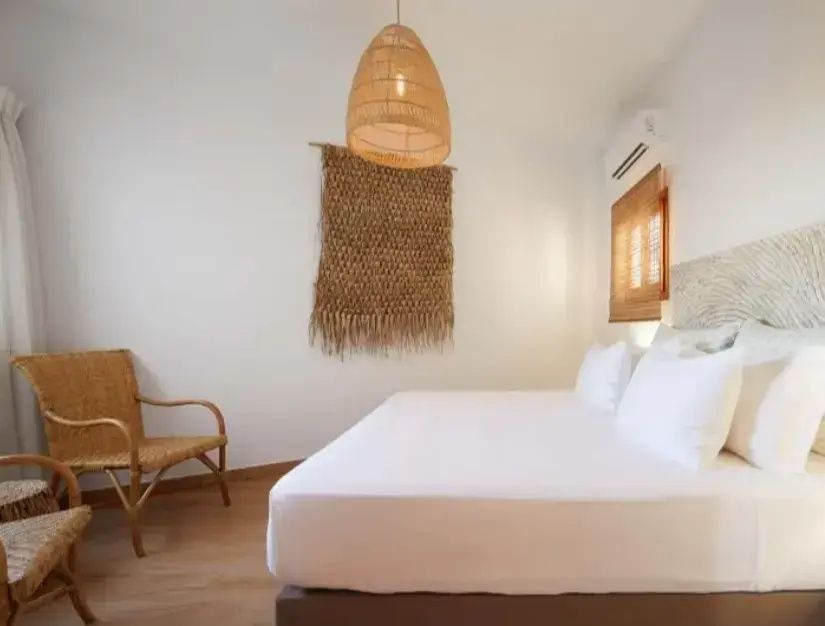 Helles Schlafzimmer mit weiß bezogenem Doppelbett, Flechtstuhl und einer Lampenaufhängung aus Rattan.