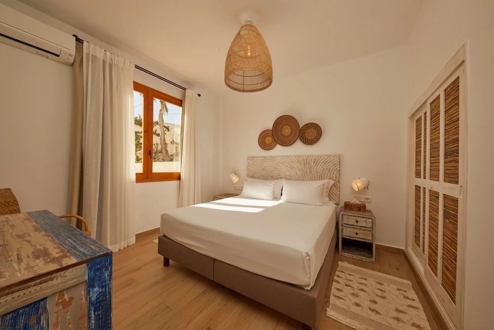 Hell eingerichtetes Schlafzimmer mit Doppelbett, Flechtlampe und Holzfenster.
