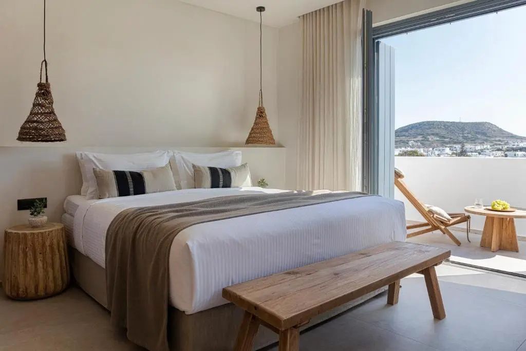 Hell eingerichtetes Doppelzimmer mit Holzbank vor dem Bett und Balkontür.