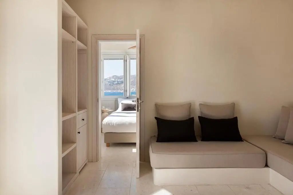 Helles Hotelzimmer mit Sitzbank, Holzregal und Blick durch eine offene Tür auf das Bett und das Meer.