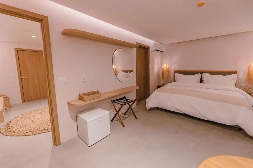 Helles Hotelzimmer mit Doppelbett, weißer Bettwäsche, Wandspiegel und Minibar.