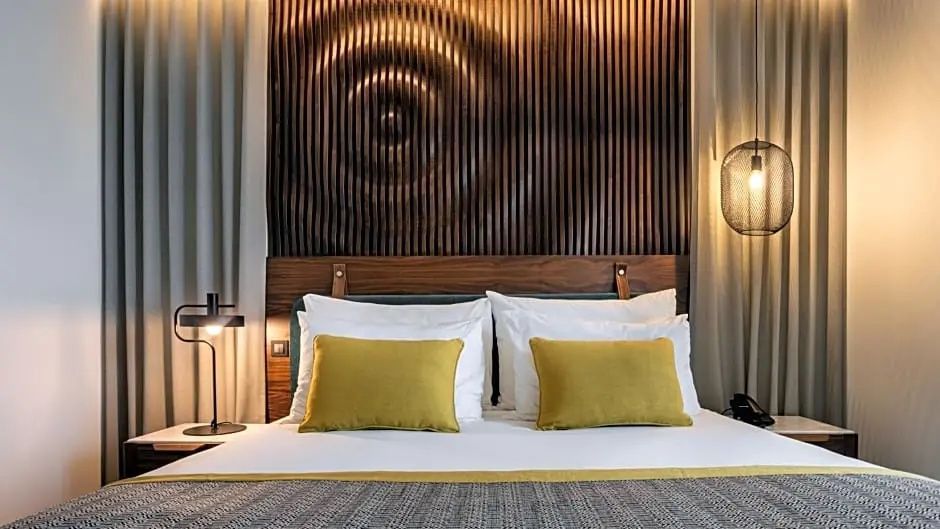 Doppelbett mit weißen Kissen, gelben Akzentkissen und einer kunstvollen Holzwand hinter dem Kopfteil.