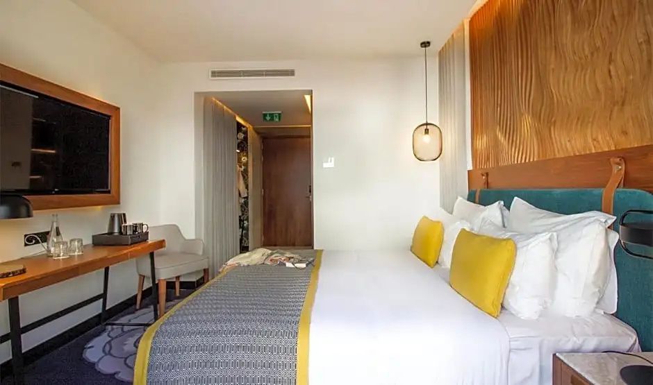 Blick über das Bett mit gelben Kissen hin zum Flur und Eingang des Hotelzimmers.