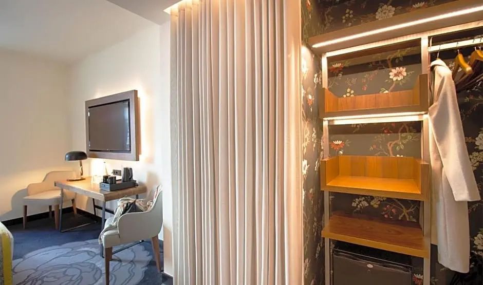 Heller Hotelraum mit offener Regaleinheit, blumiger Tapete, Vorhang und Schreibtisch mit Fernseher.