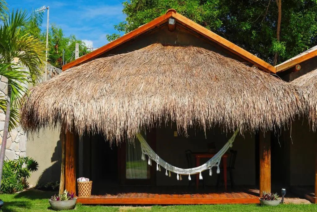 Bungalow mit Strohdach, Holzterrasse, weißer Hängematte und kleinem Rasenstück am Tag.