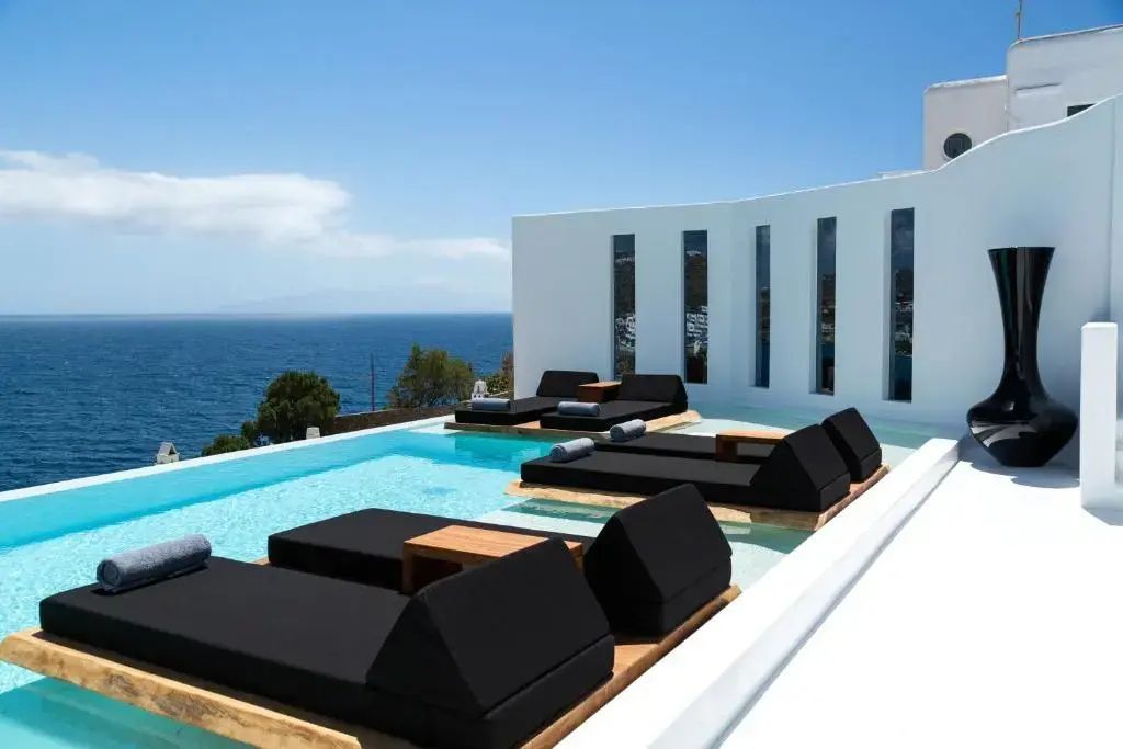 Infinitypool mit dunklen Sonnenliegen direkt im Wasser vor der Kulisse des Meeres.
