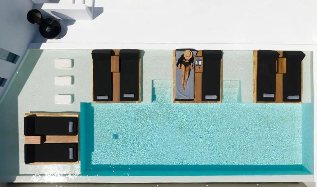 Draufsicht auf einen Pool mit dunklen Sonnenliegen im Wasser und einer liegenden Person.