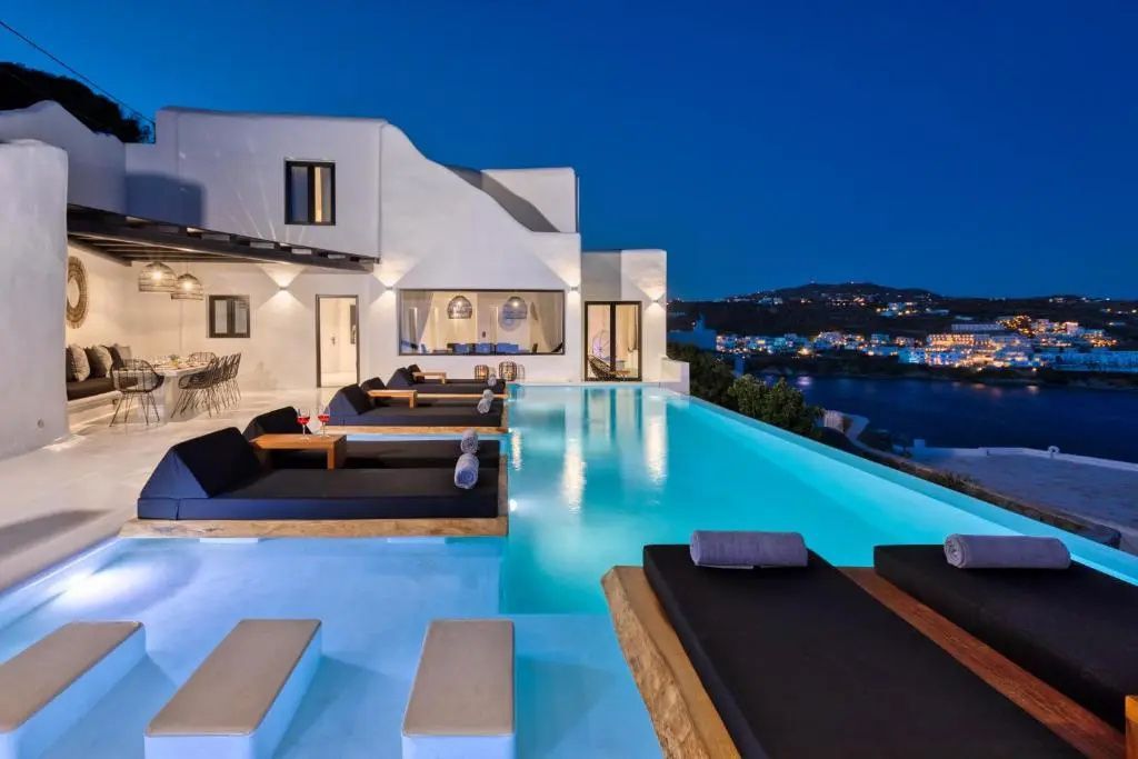 Beleuchteter Pool am Abend mit dunklen Sonnenliegen und Blick auf das Meer