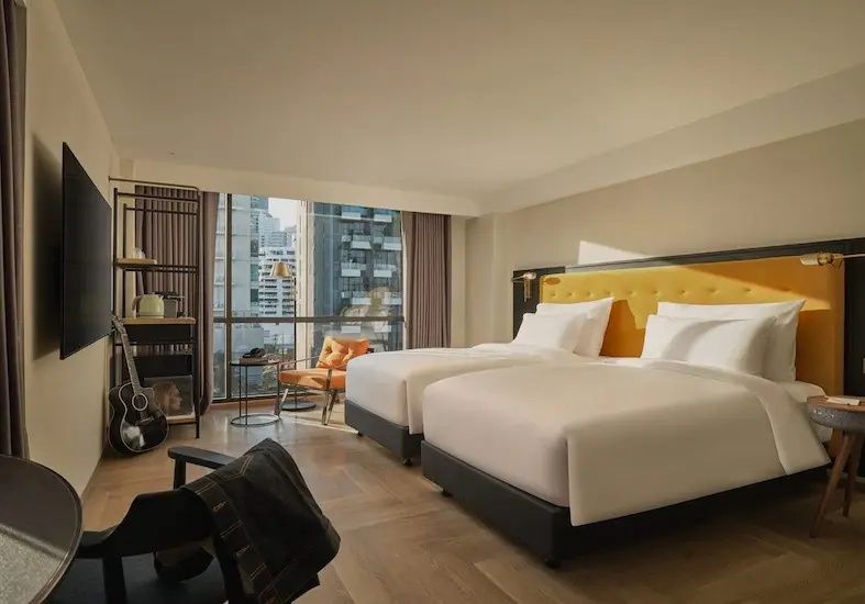 Hotelzimmer mit zwei Einzelbetten, gelbem Kopfteil und Blick auf die Stadt durch das Fenster.