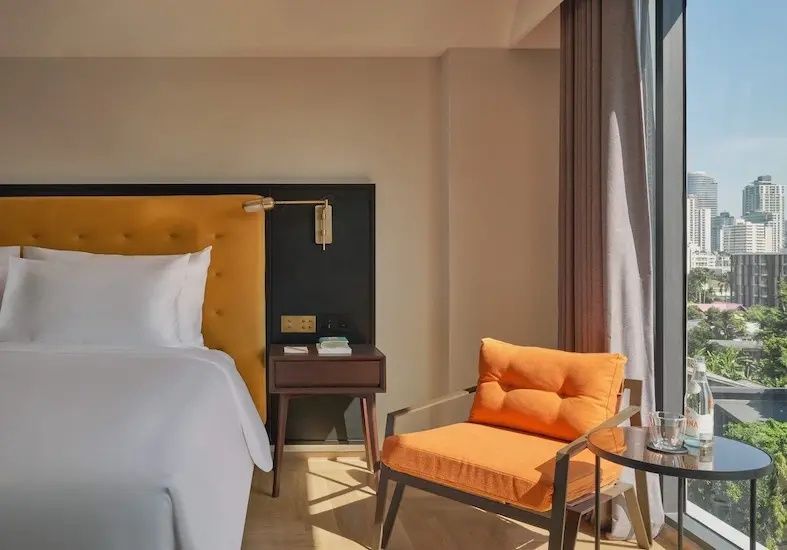 Hotelzimmer mit gelbem Kopfteil am Bett, Nachtkästchen und einem orangefarbenen Sessel.