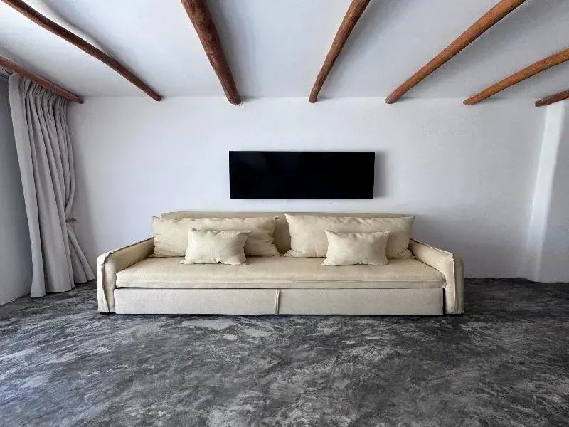 Beigefarbenes Sofa vor einer weißen Wand unter einer Holzbalkendecke mit dunklem Bildschirm.