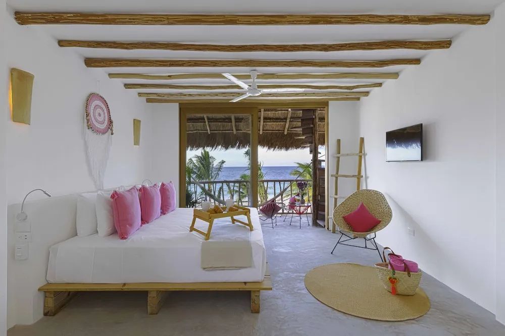 Schlafzimmer mit Holzbalkendecke, weißem Bett mit rosa Kissen und Blick auf das Meer.