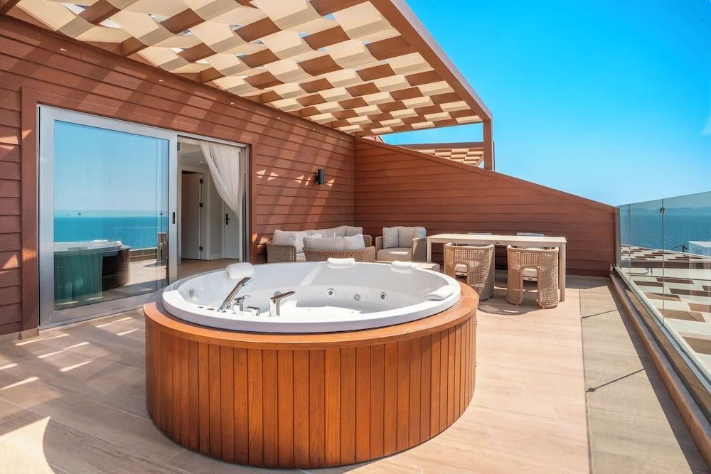 Holzterrasse mit rundem Whirlpool unter Pergola und Blick auf das blaue Meer.