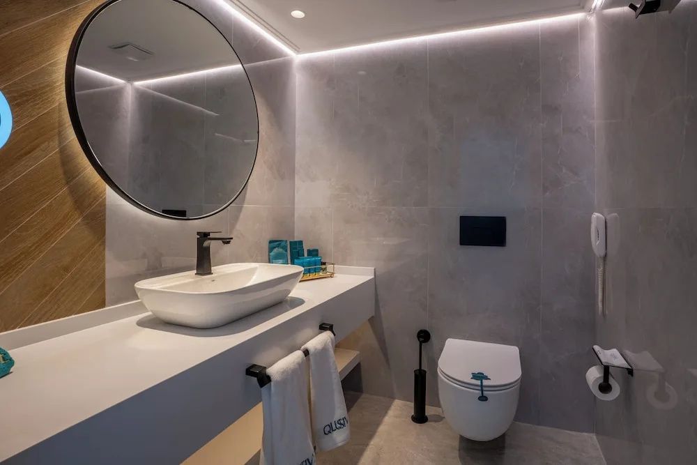 Modernes Badezimmer mit grauen Fliesen, rundem beleuchtetem Spiegel und Waschtisch.