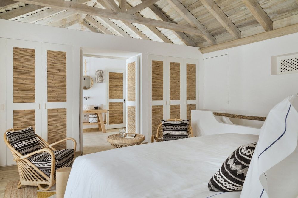 Helles Zimmer mit weiß bezogenem Bett, Sitzbereich aus Rattan und Blick in das angrenzende Badezimmer.