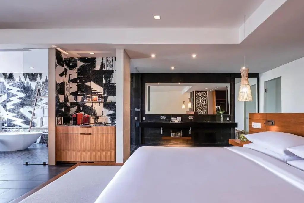 Modernes Hotelzimmer mit weiß bezogenem Bett, Minibar und schwarzem Waschtisch im Hintergrund.