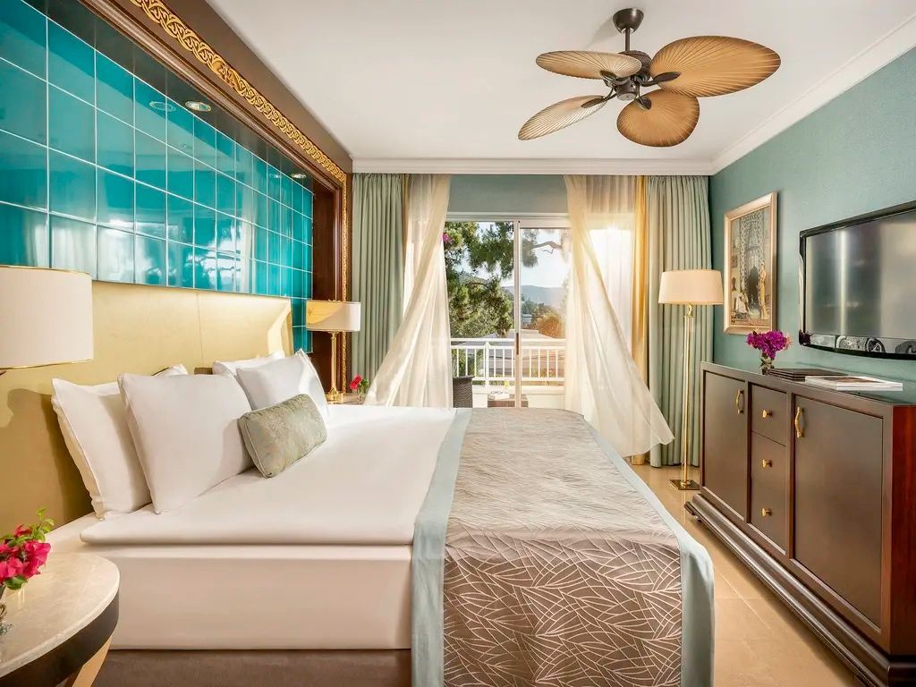 Doppelbett im Hotelzimmer mit türkisblauer Kachelwand, Deckenventilator und Balkon mit Vorhängen.