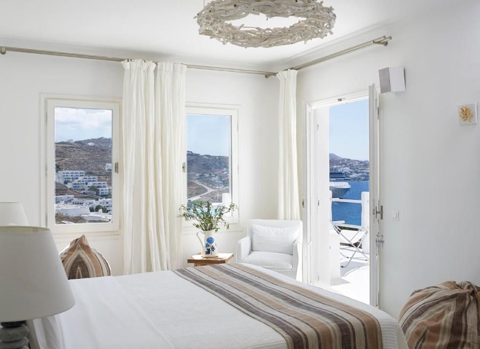 Hell eingerichtetes Schlafzimmer mit Blick durch Fenster und Balkontür auf das Meer.