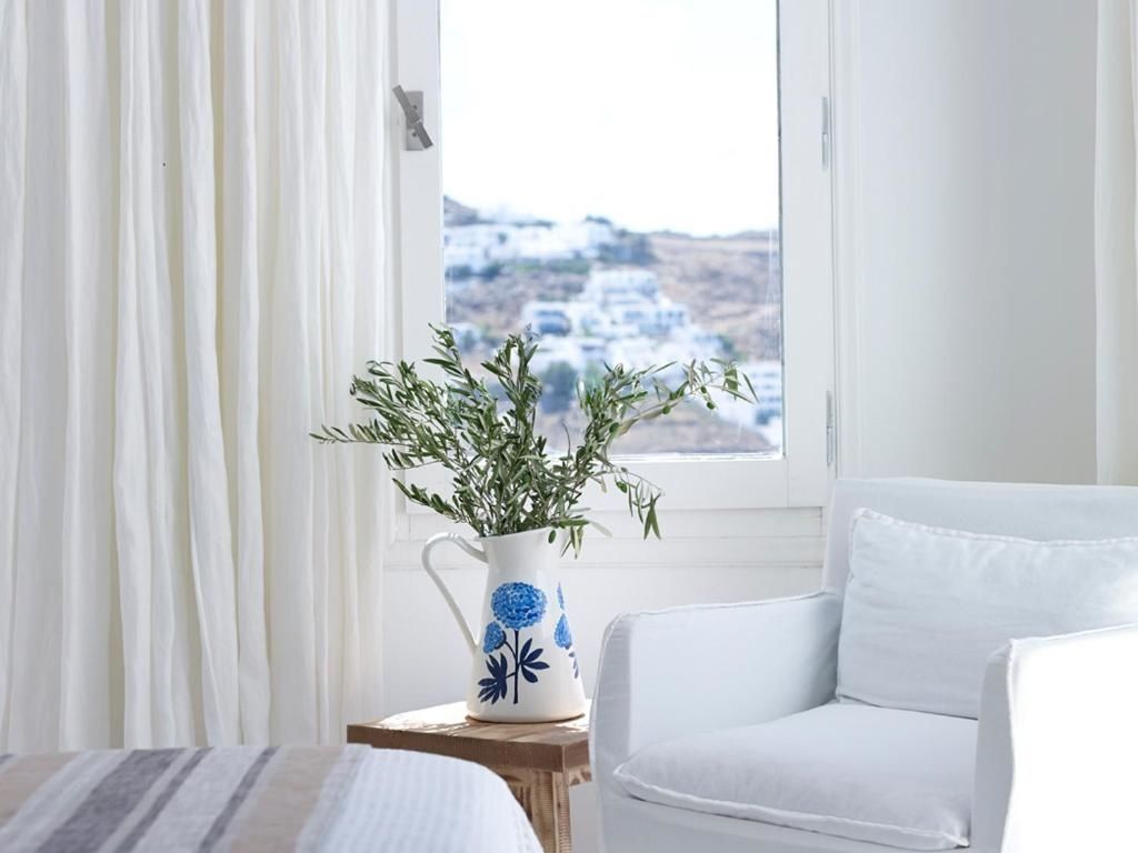 Helles Zimmer mit weißem Sessel, Vase auf Holzschemel und Fensterblick auf weiße Häuser.