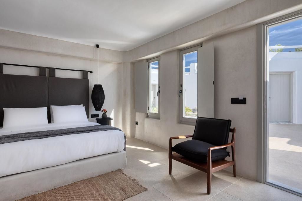 Hell eingerichtetes Schlafzimmer mit Doppelbett, schwarzem Sessel und zwei Fenstern.