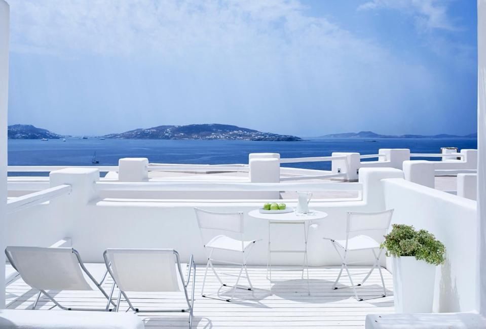 Weiße Terrasse mit Sitzgelegenheiten und Liegen bietet einen weiten Blick auf das blaue Meer und eine Insel.