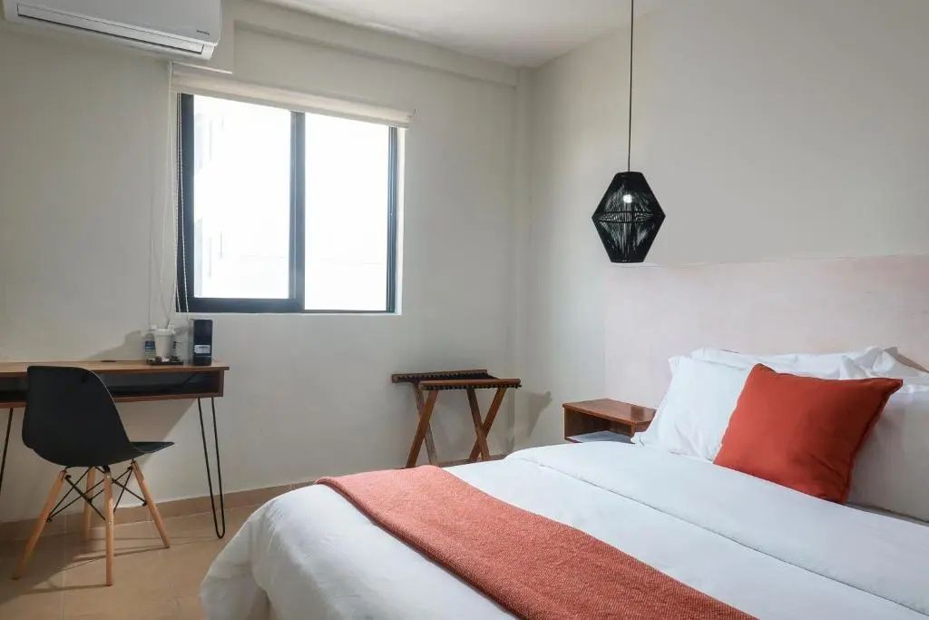 Blick im Hotelzimmer auf ein Fenster, Kofferablage und das Bett mit oranger Decke.