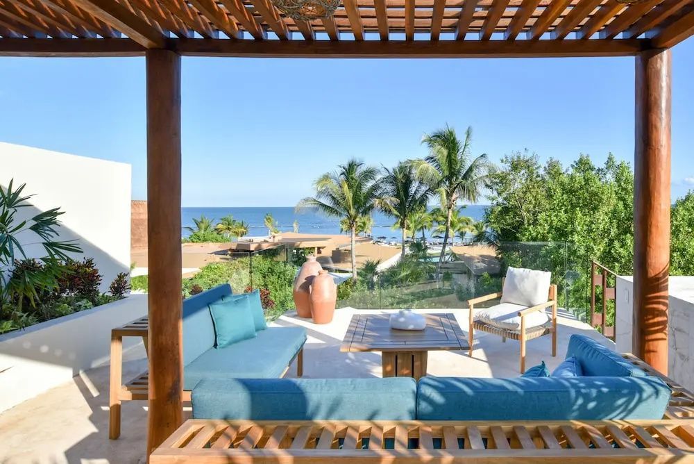 Überdachte Terrasse mit blauen Polstermöbeln und Ausblick auf Palmen sowie das Meer.