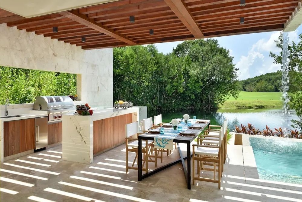 Überdachter Essbereich auf einer Terrasse mit Esstisch, Grillstation und Blick auf das Wasser.