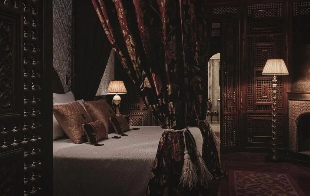 Doppelbett mit braunen Kissen, beleuchteter Nachttischlampe und dunklen Vorhängen mit Quasten.