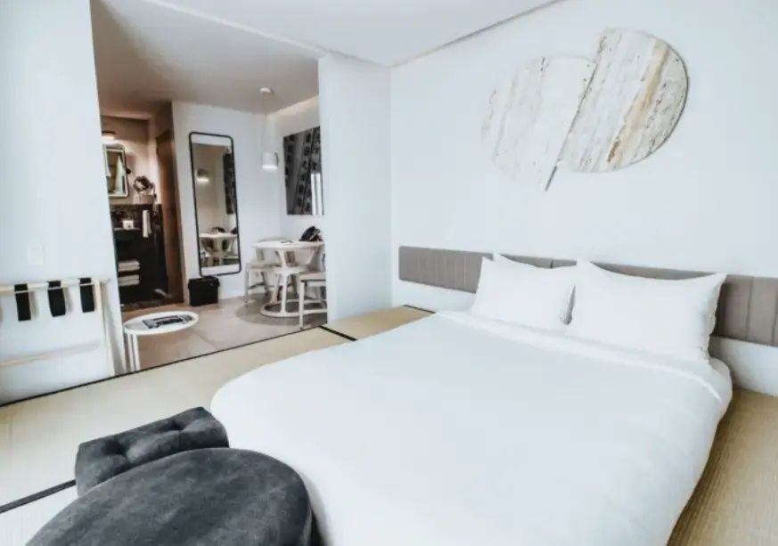 Hotelzimmer mit weißem Bett auf Tatamimatten und Blick auf den Sitzbereich im Hintergrund.