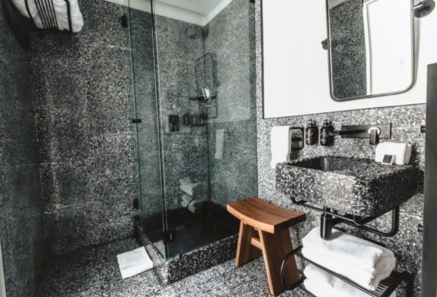 Badezimmer mit dunklen Granitwänden, einer Glasdusche, Waschtisch und einem Holzschemel.