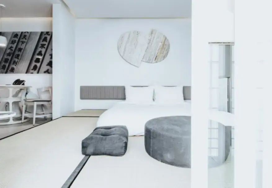 Helles Hotelzimmer im japanischen Stil mit Tatamiboden, weißem Bett und grauem Sitzpuff.