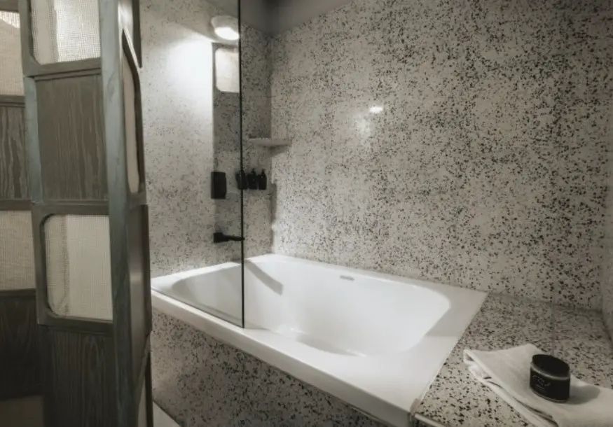 Weiße Badewanne mit Duschglaswand und hellem Terrazzomuster an der Wand.