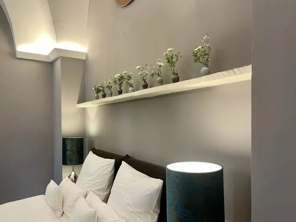 Bett mit weißen Kissen, Nachttischlampe und schmaler Wandablage mit kleinen Blumenvasen.