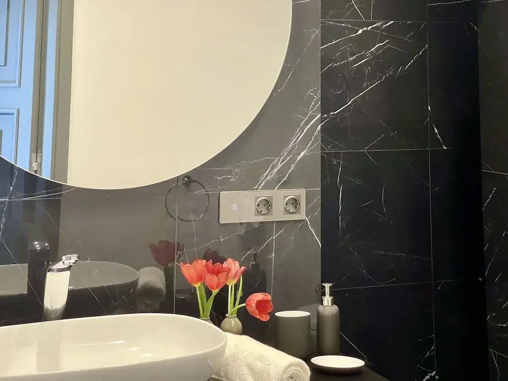 Waschtisch im Badezimmer mit rundem Spiegel, dunklen Marmorfliesen und frischen Tulpen.