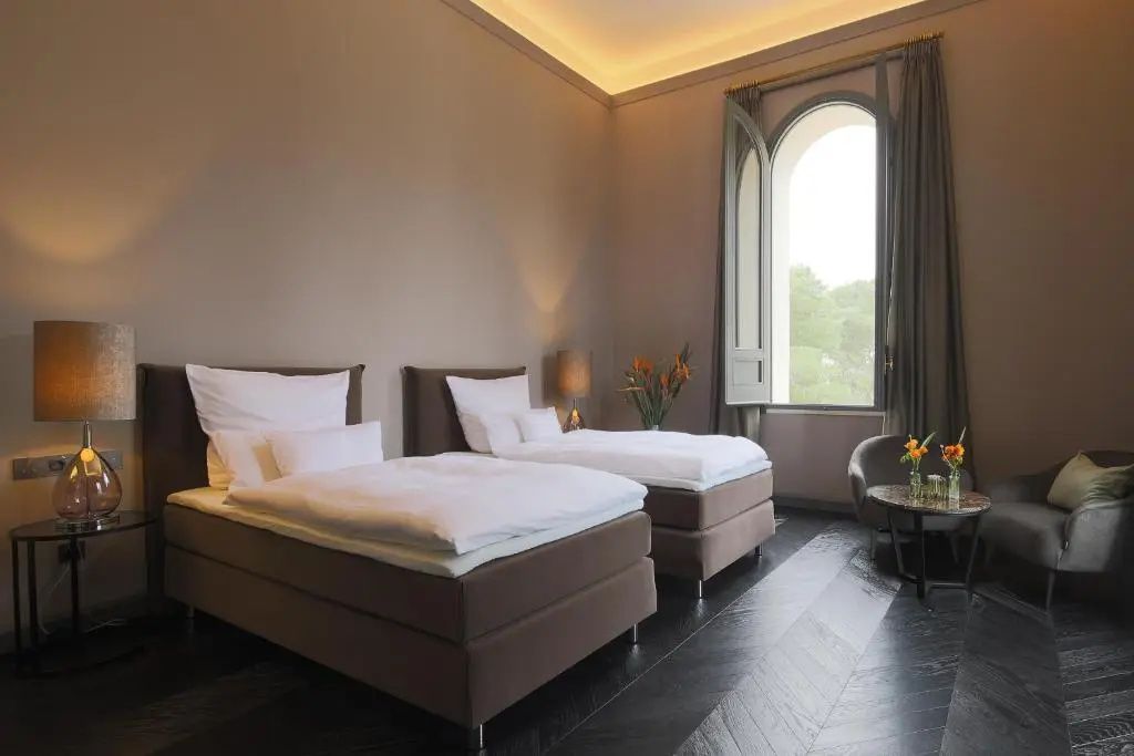 Zwei getrennte Betten in einem Hotelzimmer mit hoher Decke und Bogenfenster.