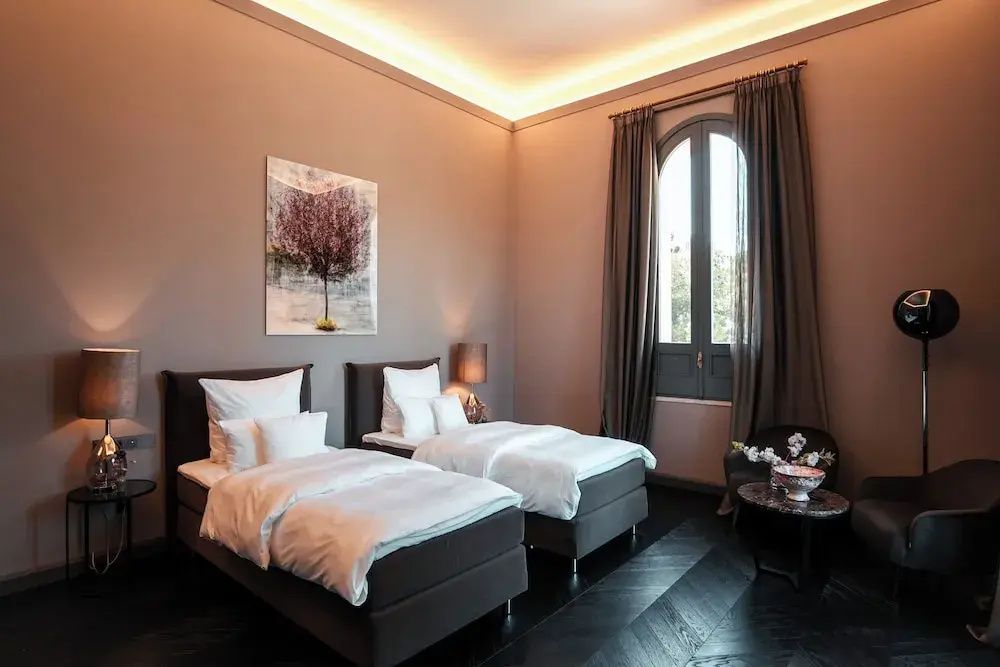 Schlafzimmer mit zwei Einzelbetten, dunklen Vorhängen und einem hohen Rundbogenfenster.