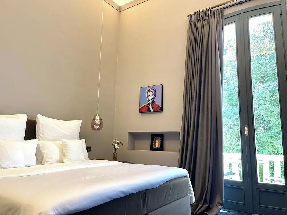 Doppelbett in hellem Zimmer mit bodentiefem Fenster, Vorhang und Bild in einer Wandnische.